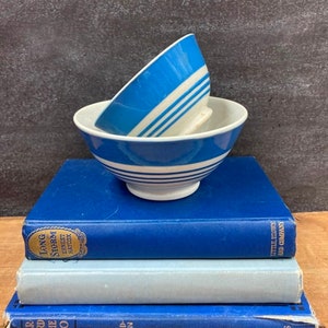 May include: Two blue and white striped bowls stacked on top of three vintage books. The books are blue and light blue, with titles and author names visible. The bowls have a classic design.