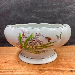 May include: White ceramic bowl with a scalloped rim, featuring a floral and butterfly design. The bowl has a pedestal base and is decorated with green, pink, and brown accents. A decorative piece for the home.