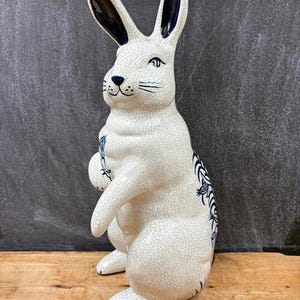 May include: A ceramic rabbit figurine with a white crackle glaze finish. The rabbit has black and blue accents on the ears and decorative patterns on the body. The figurine is standing upright on its hind legs.