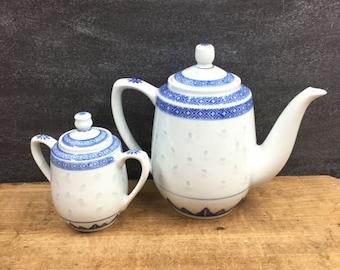 May include: A white porcelain teapot and creamer set with blue floral patterns. The teapot has a long spout and a curved handle. The creamer has two handles. Both pieces have matching lids with small knobs. The set is on a wooden surface.