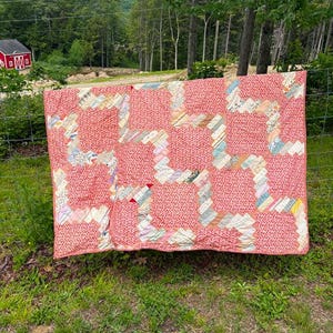 May include: A rectangular quilt featuring a repeating pattern of large coral-red fabric squares interspersed with diagonal strips of multi-colored, patterned fabric. The quilt displays a complex, interlocking geometric design throughout its surface.