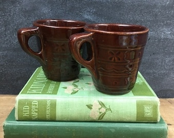 May include: Two brown ceramic mugs with a brown glaze and a decorative pattern. The mugs are sitting on top of a stack of two green books. The top book has the title "Kidnapped" and the author "Robert Louis Stevenson".