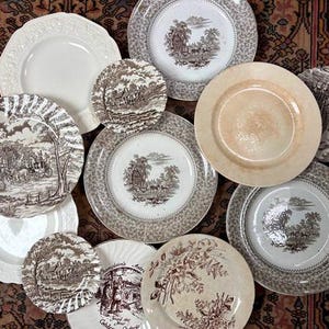 May include: A collection of eleven ceramic plates featuring a mix of solid cream-colored pieces and brown transferware designs. The patterned plates display detailed landscape scenes, including horse-drawn carriages, rural buildings, and floral motifs.