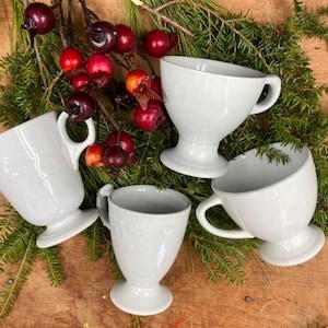 May include: Four white ceramic cups with handles and a pedestal base. The cups are arranged on a wooden surface with green foliage and red berries.