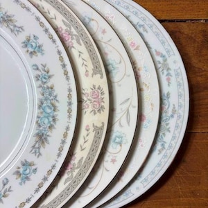 May include: A stack of decorative ceramic plates with floral patterns. The plates feature various designs, including blue, pink, and yellow flowers, with gold trim. The plates are arranged on a wooden surface.
