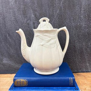 May include: Cream-colored ceramic teapot with a wheat stalk design, lid, and curved handle. The teapot sits atop two blue hardcover books. The books have gold lettering and are stacked on a wooden surface.