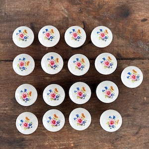 May include: A collection of white ceramic cabinet knobs with floral designs in pink, orange, and blue. The knobs are round and arranged on a wooden surface. The floral pattern adds a touch of vintage charm.