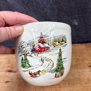 May include: A white ceramic mug with a winter scene featuring a red church, snow-covered trees, and a horse-drawn sleigh. The mug has a curved shape and a smooth surface, perfect for hot beverages.