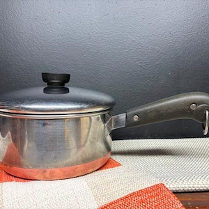 May include: A stainless steel saucepan with a lid and a dark handle. The pot has a copper-colored bottom and sits on a patterned fabric. The handle has a loop at the end. The lid has a black knob.