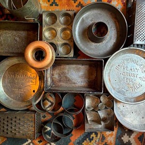 May include: An assortment of vintage baking tools, including cake pans, pie plates, muffin tins, and cookie cutters. The items are made of metal and show signs of wear, with a rustic aesthetic. The arrangement is on a patterned rug.