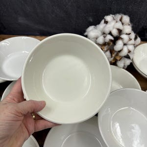 May include: A collection of off-white ceramic bowls and plates. The bowls have a rounded shape with a slightly raised rim. The plates are flat with a subtle edge. A cotton branch is in the background.