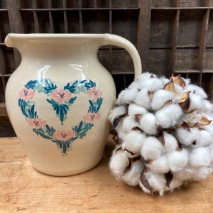 May include: A cream-colored ceramic pitcher with a heart-shaped floral design in pink and teal. Beside it is a fluffy cotton ball arrangement with brown accents, all set on a wooden surface.
