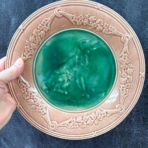 May include: A round ceramic plate featuring a central emerald green circular panel with a raised relief of a dog. The outer rim is a muted dusty pink color, decorated with a continuous embossed floral garland pattern and tied ribbon motifs.