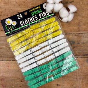May include: A package of 24 rustproof spring clothes pins in yellow, white, and green. The package has a black label with white text and a daisy graphic. The package is on a wooden surface.