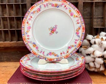 May include: Set of four vintage floral patterned plates with a red and pink floral border. The plates are stacked on top of each other and are sitting on a book.