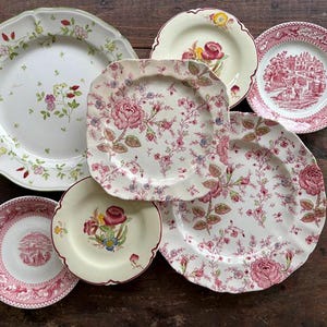 May include: A collection of vintage floral patterned plates in various shapes and sizes. The plates feature pink and red roses, green leaves, and other floral designs on a white background. Some plates have scalloped edges.