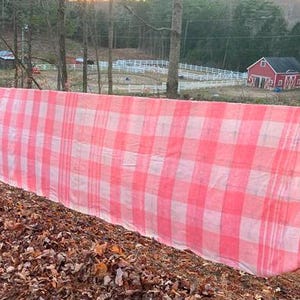May include: A large pink and white checkered tablecloth is draped over a fence. The tablecloth is made of a lightweight fabric and has a faded appearance.