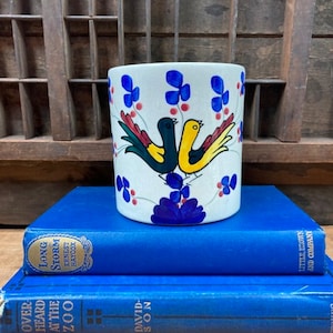 May include: A white ceramic planter with a blue, yellow, and green floral design and two birds. The birds are facing each other and are painted in black, yellow, and red. The planter is sitting on top of two blue books.