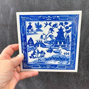May include: A square ceramic tile featuring a blue and white Willow pattern. The design depicts a landscape with a pagoda, a bridge, a boat, and trees, all enclosed within a decorative geometric border. The tile has a smooth, glossy finish.