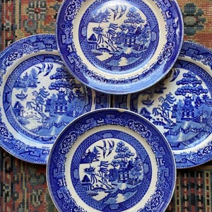 May include: Four blue and white porcelain plates with a traditional Chinese willow pattern. The plates have a white background with a blue willow tree, a bridge, and a pagoda.
