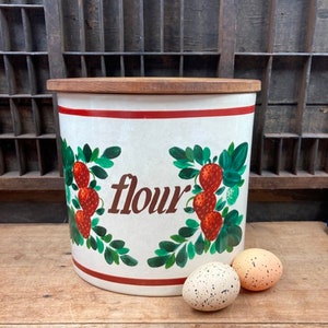 May include: A white ceramic flour canister with a wooden lid, adorned with red strawberries and green leaves. The word "flour" is written in brown script. Two speckled eggs sit in front of the canister.