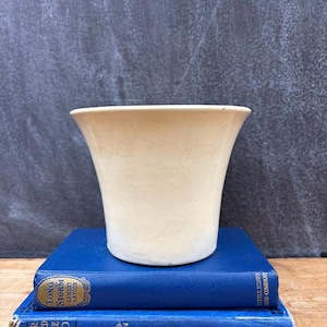 May include: A cream-colored, flared ceramic vase sits atop two vintage blue books. The vase has a smooth, matte finish. The books have gold lettering and are stacked on a wooden surface.