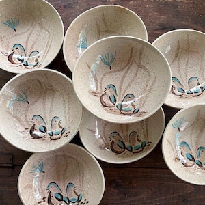 May include: A set of seven round ceramic bowls with a speckled cream finish. Each bowl features a hand-painted design of two small blue and brown birds perched near a stylized plant, with a light brown brushstroke pattern in the background.