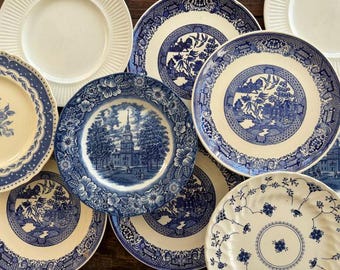 May include: A collection of vintage ceramic plates with blue and white floral and landscape designs. Some plates have scalloped edges, while others feature intricate patterns. The plates vary in size and style, creating a charming, eclectic display.