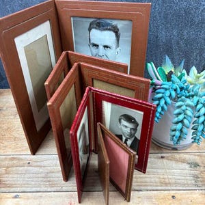 May include: A collection of vintage brown and red photo frames, some with black and white photographs. The frames are made of a leather-like material and are arranged on a wooden surface. A small succulent plant is in the background.