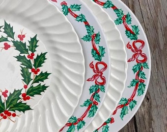 May include: A stack of white ceramic plates with a festive Christmas design. Each plate features a holly and berry wreath and a red ribbon border. The plates have a fluted edge.