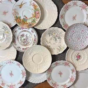 May include: A collection of vintage porcelain plates and saucers. The plates feature floral patterns, intricate designs, and varying edge details. Colors include white, pink, green, and gold. Some plates have embossed patterns, while others showcase detailed illustrations of birds and flowers.