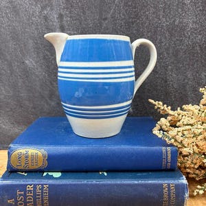 May include: A ceramic pitcher featuring a white base with horizontal blue stripes of varying widths. The pitcher has a curved handle on the right side and a pouring spout on the left. The design is consistent around the body of the vessel.
