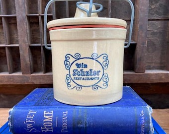 May include: A white ceramic crock with a blue and white logo that reads "Win Schuler Restaurants". The crock has a metal wire lid and is sitting on top of two blue books.