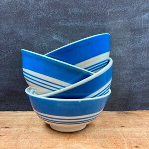 May include: A stack of four ceramic bowls. Each bowl is blue with white stripes and a white rim. The bowls are stacked on top of each other, with the top bowl slightly tilted. The background is a dark gray.