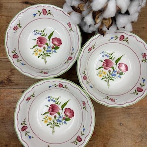 May include: Three white ceramic bowls with scalloped edges and a green trim. Each bowl features a floral design with red, pink, yellow, and blue flowers. The bowls are arranged on a wooden surface.
