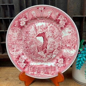 May include: A pink and white ceramic plate with a red and white design of the Natural Bridge of Virginia. The plate features images of the bridge, surrounding trees, and two men in portraits. The text "Natural Bridge of Virginia" is printed on the plate.