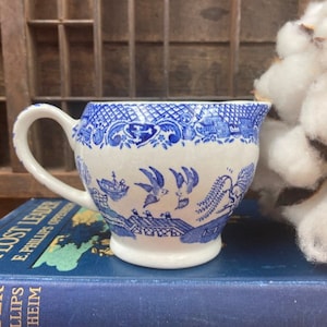 May include: A blue and white ceramic creamer with a floral design and a bird motif. The creamer is sitting on a blue book.