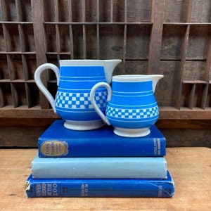 May include: Two blue and white ceramic pitchers with checkered patterns. The larger pitcher has a handle and a spout. The smaller pitcher has a handle. Both pitchers are sitting on a stack of blue books.