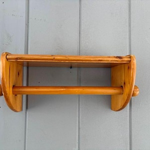 May include: A wooden paper towel holder with a shelf. The holder is made of light-colored wood and has a horizontal dowel for holding the paper towels. The shelf is above the dowel. The holder is mounted on a gray wall.