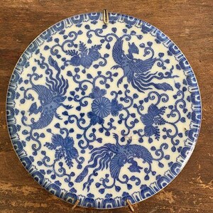 May include: Round, white and blue decorative plate with a phoenix and floral design. The plate has a blue border and is supported by a gold metal stand. The plate's design features blue phoenixes and floral patterns.