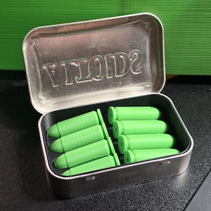 May include: A rectangular silver metal tin with an open lid featuring the embossed text "VOID". Inside the tin are eight bright green, bullet-shaped objects arranged in two rows, held in place by a custom-fitted black plastic insert.