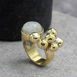 May include: A gold-toned ring featuring a smooth, round, translucent white stone set on one side, complemented by a cluster of five polished gold-toned spheres of varying sizes attached to the band next to the stone.