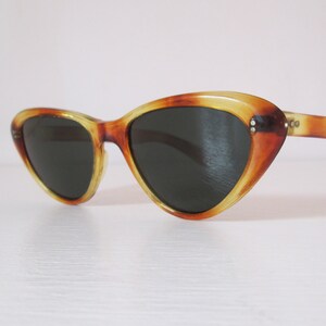 Puede incluir: Gafas de sol de ojo de gato con estampado de carey. Las monturas son una mezcla de marr&oacute;n y amarillo, con lentes oscuras. Las gafas de sol tienen un estilo retro y vintage, sobre un fondo blanco.