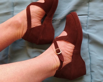 May include: A pair of brown suede wedge shoes with ankle straps and a buckle closure. The shoes are on a light blue surface.
