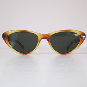 Puede incluir: Gafas de sol de ojo de gato con montura con estampado de carey. Las gafas de sol tienen lentes oscuras y un dise&ntilde;o retro cl&aacute;sico. La montura es una mezcla de tonos &aacute;mbar y marr&oacute;n, con una ligera curva ascendente en los bordes exteriores.