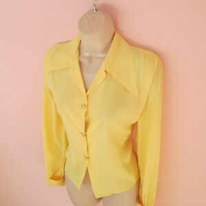 May include: A pale yellow long-sleeved blouse with a classic collar and button-down front. The blouse is displayed on a mannequin, showcasing its tailored fit and design. The cuffs are buttoned, and the fabric appears to be lightweight.