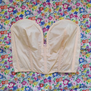 Pode incluir: Um bustiê pêssego claro com decote em V profundo. O corpete tem costuras verticais e um design estruturado. A peça é exibida em um fundo de tecido com estampa floral.
