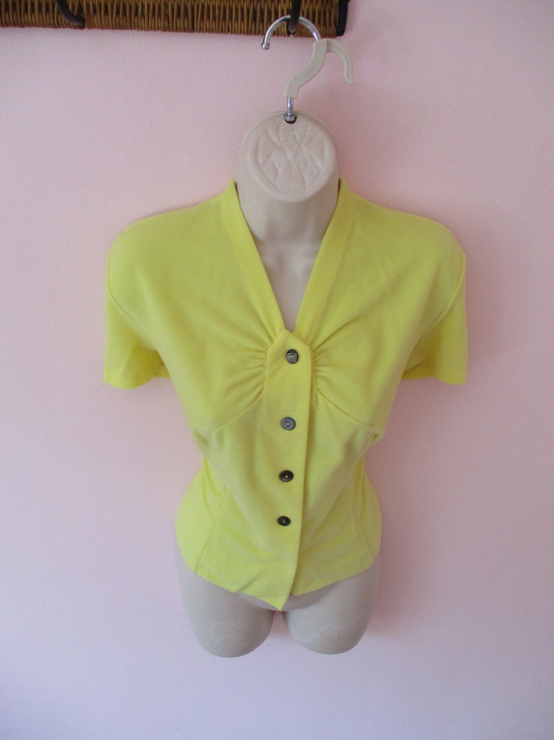 K&ouml;nnte beinhalten: Eine leuchtend gelbe Bluse mit kurzen &Auml;rmeln, V-Ausschnitt und Knopfleiste. Die Bluse wird auf einer Schaufensterpuppe pr&auml;sentiert und hat eine taillierte Taille. Die Kn&ouml;pfe sind dunkelgrau. Der Hintergrund ist hellrosa.