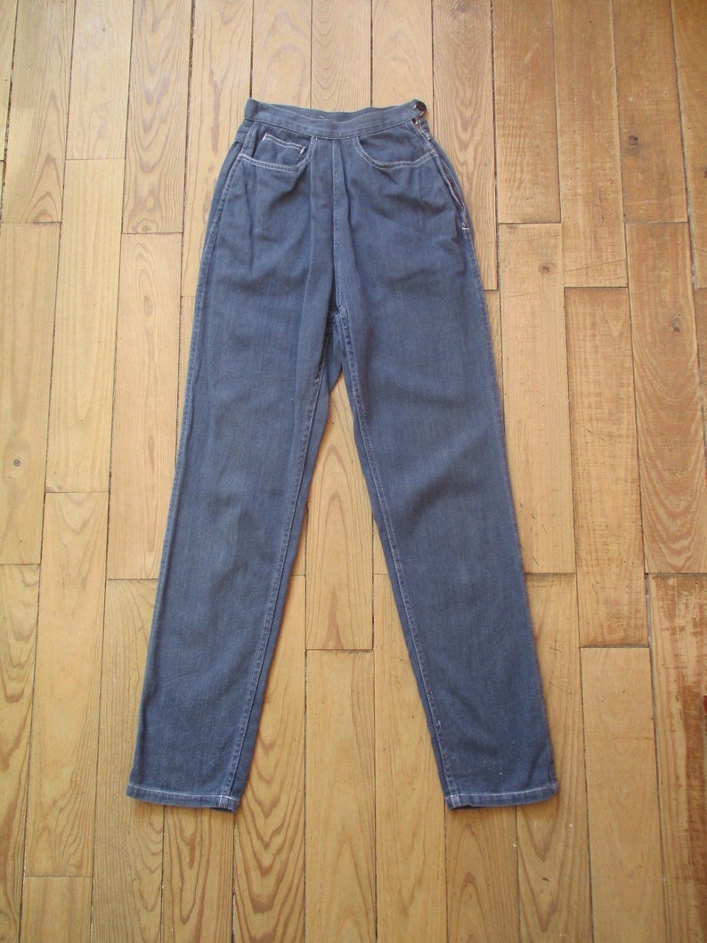 May include: Dark blue denim pants with a high waist and straight leg. The pants have two front pockets and a button and zipper closure. The stitching is white. The pants are displayed on a wooden surface.