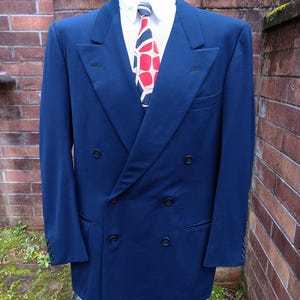 Puede incluir: Una americana azul marino de doble botonadura con solapas de pico y seis botones frontales, sobre una camisa blanca de cuello rígido y una corbata con un llamativo patrón geométrico en rojo, azul marino y crema, expuesta en un maniquí.
